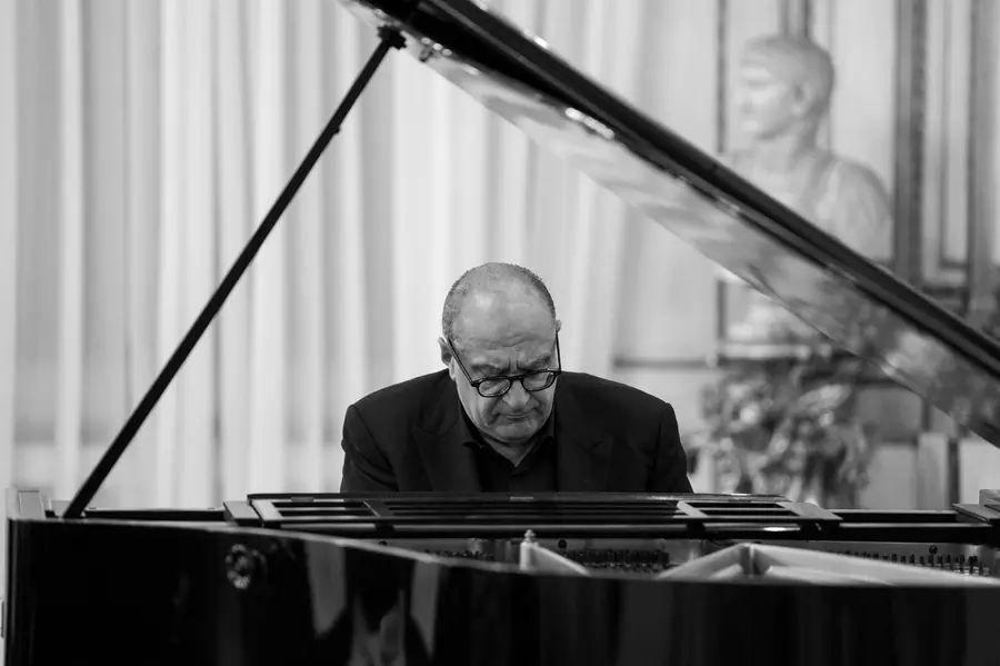 In bianco e nero, seduto al pianoforte a coda con lo sguardo basso sulla tastiera; sullo sfondo un busto di marmo e una tenda chiara