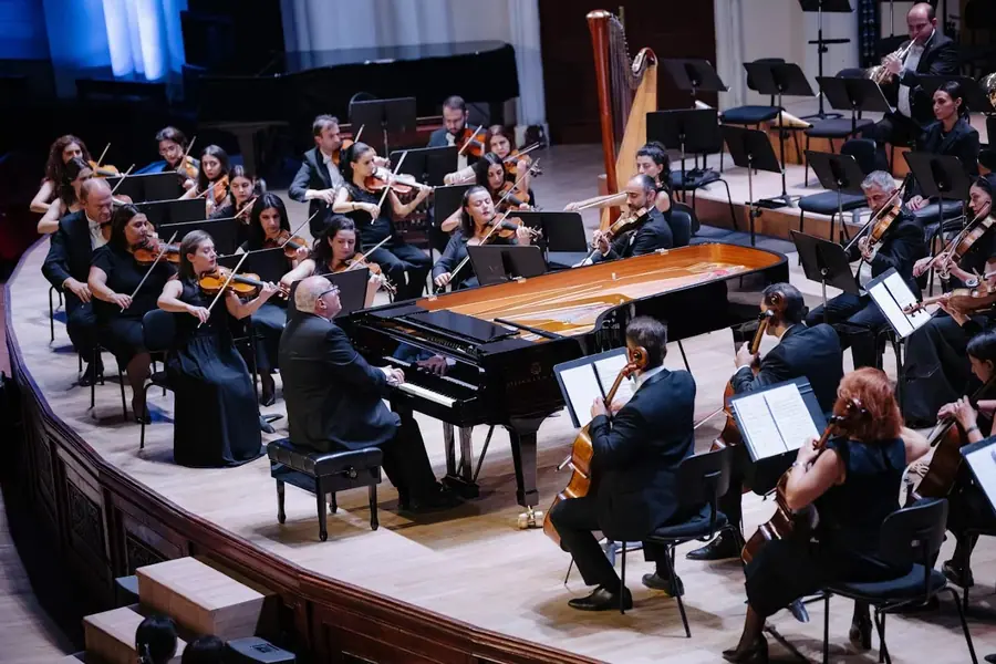 Il pianista al gran coda aperto al centro dell'orchestra, ripreso dall'alto: archi a sinistra, violoncelli e arpa a destra, luce calda sul legno del pianoforte.