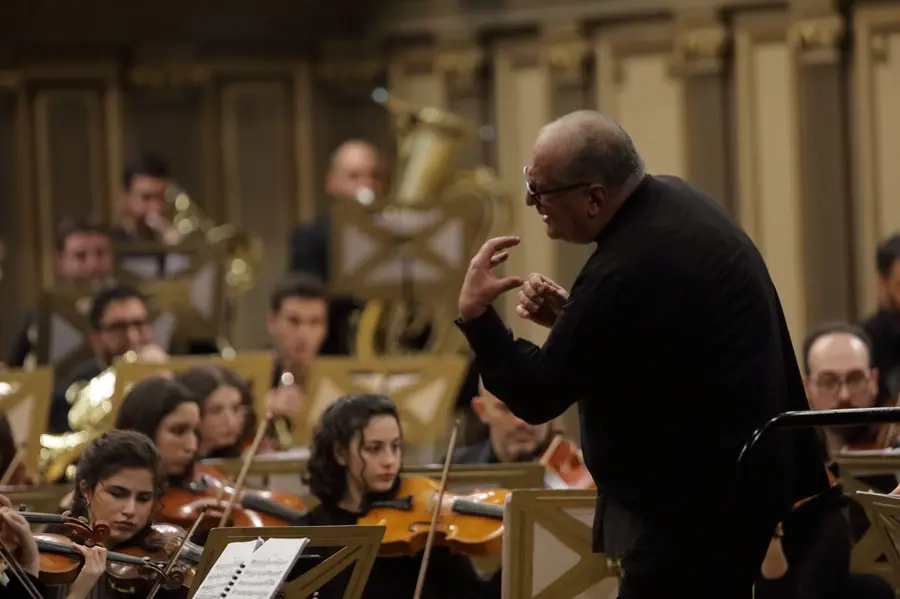 Three-quarters from behind, he conducts with bare hands under a warm golden light, before a young orchestra of strings and brass in an ornately decorated hall