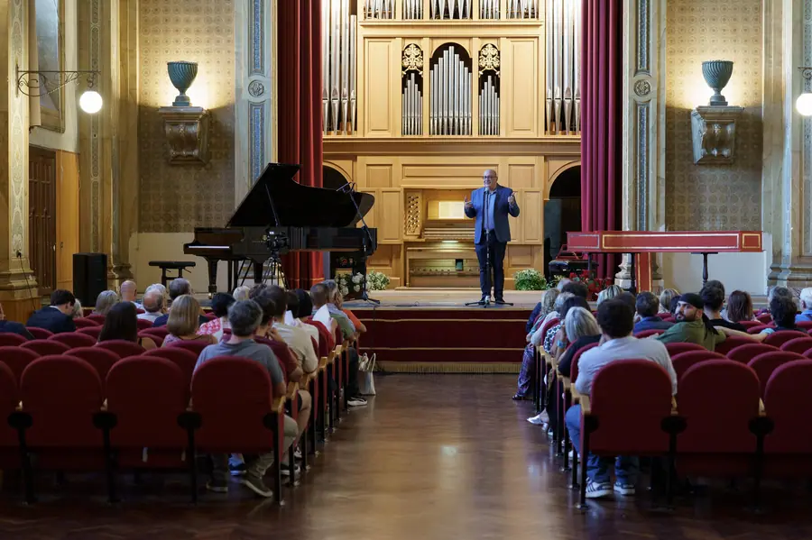 Veduta d'insieme di una sala con grande organo a canne, pianoforte a coda a sinistra e clavicembalo rosso a destra; un uomo parla al microfono sul palco davanti al pubblico in poltrone rosse.