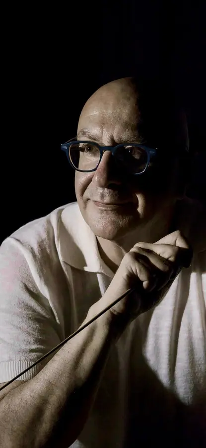Close portrait in half-light: a man with blue glasses and a pale polo shirt, the baton held between his hands