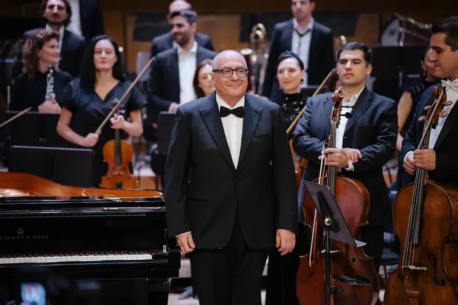 In dinner jacket and bow tie, standing beside a Steinway grand piano, he smiles towards the audience while the orchestra applauds behind him