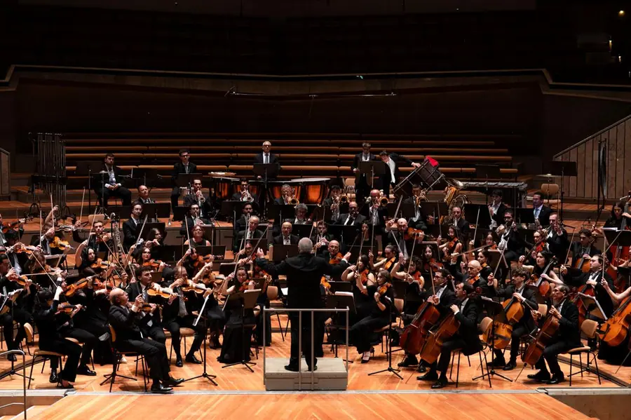 Full symphony orchestra on a pale wooden stage in a modern terraced hall; the conductor stands with his back to us on the rostrum, timpani and brass at the rear.