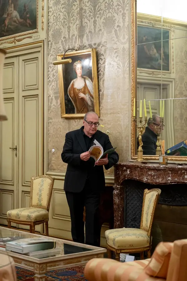 Stessa scena della precedente, ripresa un poco piu larga: in piedi davanti al camino con il programma in mano, fra le sedie dorate del salotto