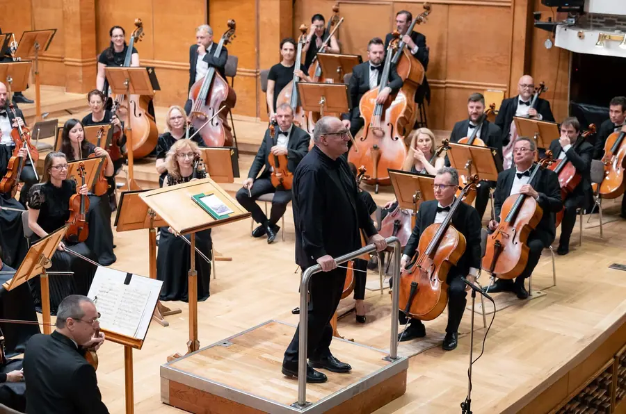 On the raised podium at the centre of the stage, turned towards the hall, with the full symphony orchestra set around him in a hall with pale wooden walls