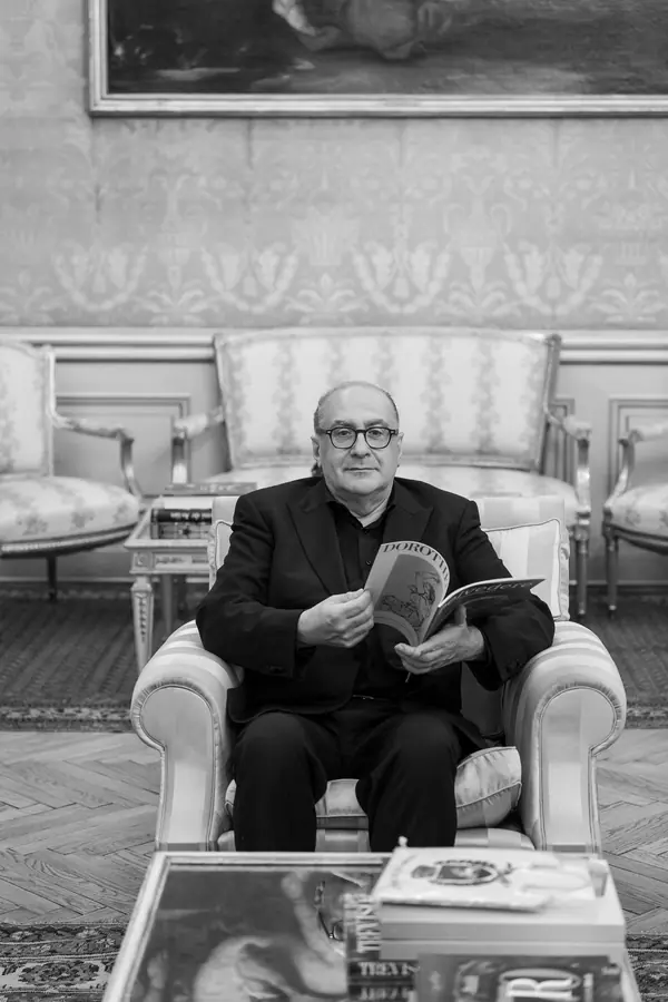 In black and white, seated in an armchair in a period drawing room with a magazine in his hands, books on the low table in the foreground