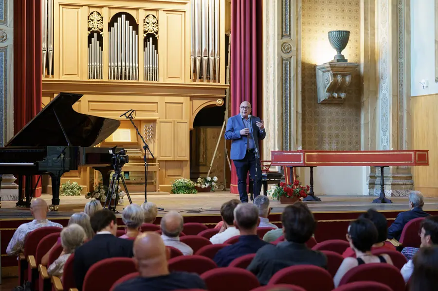 The stage seen from the stalls: the man in a blue jacket at the microphone between the grand piano, the pipe organ and the harpsichord, with the heads of the audience in the foreground.