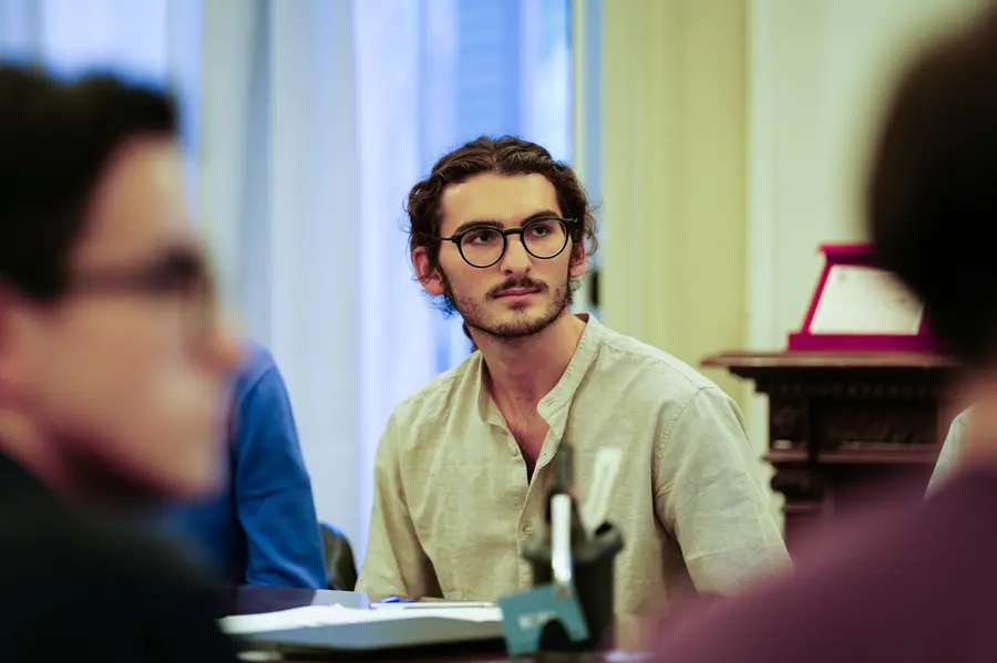 Close-up of a young man with curly hair, round glasses and a pale linen shirt, seated and attentive; blurred figures in the foreground
