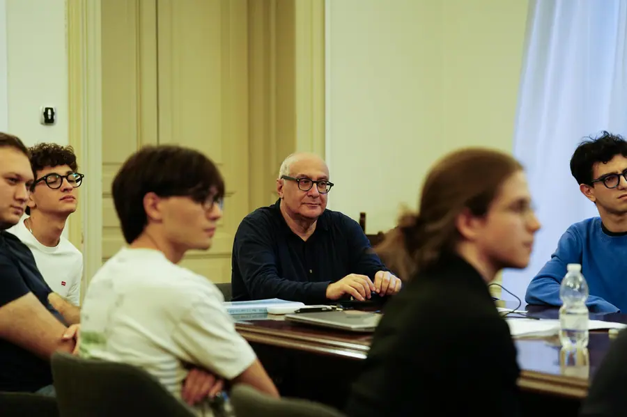 A room with a high ceiling: the bald man with glasses sits at the centre behind a dark table, surrounded by young people blurred in the foreground