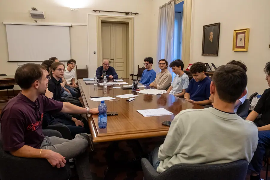 Sala con schermo di proiezione e ritratti alle pareti: l'uomo con occhiali siede in fondo al lungo tavolo, giovani seduti su entrambi i lati con fogli davanti
