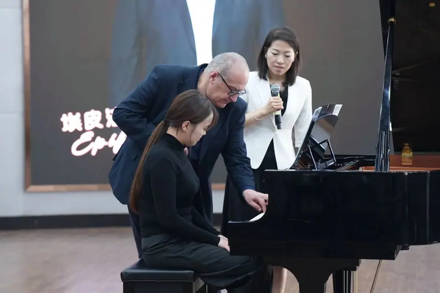 Bent over the keyboard beside a young female student seated at the grand piano, while a woman with a microphone translates next to them