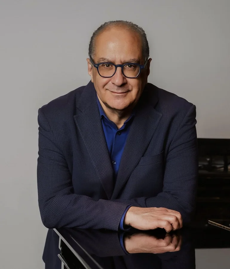 Colour portrait: in a dark blue jacket and blue shirt, leaning on the lid of a black grand piano against a light grey background
